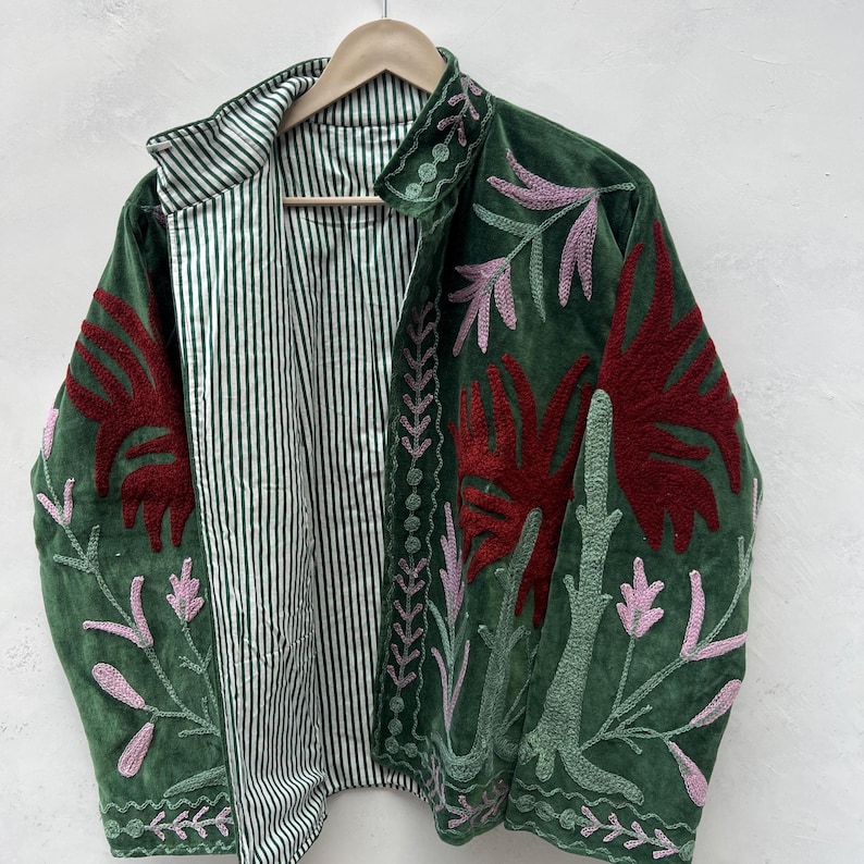 May include: A green velvet jacket with embroidered floral designs in red, pink, and green. The jacket has a striped lining and a collared neckline. The jacket is hanging on a wooden hanger.