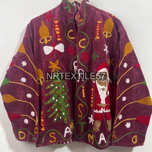 May include: Burgundy velvet jacket with embroidered Christmas-themed designs. Features a Christmas tree, Santa Claus, bells, and the word "CHRISTMAS" in white. The jacket is hanging on a wooden hanger.