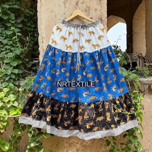 Könnte beinhalten: Ein mehrlagiger Rock mit weißem, blauem und schwarzem Design. Der Rock hat einen sich wiederholenden Tigerdruck in Gold. Der Text "NRTEXTILE" ist auf der blauen Ebene aufgedruckt. Der Rock hängt an einem Holzbügel.