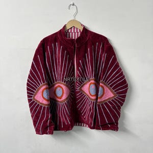 Könnte beinhalten: Burgunderfarbene Samt-Reißverschlussjacke mit gestickten Augendesigns. Die Jacke hat einen hohen Kragen, ein gestreiftes Futter und den Text "NRTEXTILE". Die Augen sind rosa, blau und braun, mit weißen Strahlenlinien.