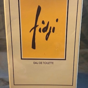 May include: A rectangular box of "Fidji" Eau de Toilette by Guy Laroche Paris. The box has a cream-coloured background with a yellow square in the centre. The word "Fidji" is written in black script. The text "EAU DE TOILETTE" is below the script.