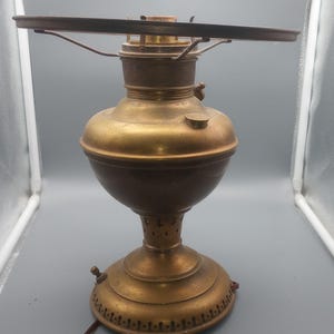 May include: A vintage-style brass table lamp with a rounded central reservoir, a wide circular top rim, and a flared base featuring decorative cutouts. The lamp has a metallic finish and includes a brown electrical cord extending from the side.