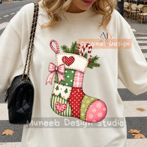May include: A cream-colored crewneck sweatshirt featuring a large graphic of a patchwork Christmas stocking. The stocking is decorated with red and green patterns, including polka dots, stripes, and snowflakes, accented by a pink bow and hearts.