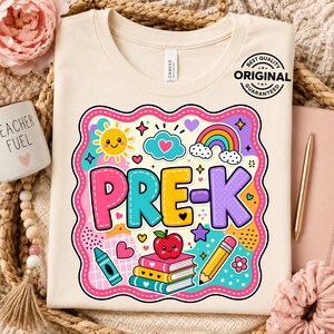 May include: A cream-colored t-shirt featuring a colorful graphic with the text PRE-K. The design includes a smiling sun, a rainbow, a stack of books with a red apple on top, a pencil, and a crayon, all enclosed within a scalloped pink border.