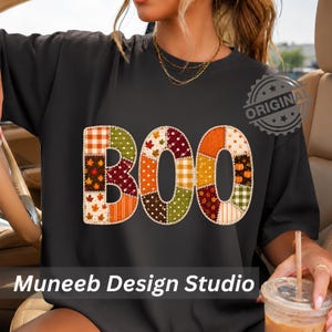 May include: A black t-shirt featuring the word BOO in large, block letters. Each letter is composed of a patchwork design with various autumn-themed patterns including orange plaid, brown leaves, white polka dots, and small orange pumpkins.