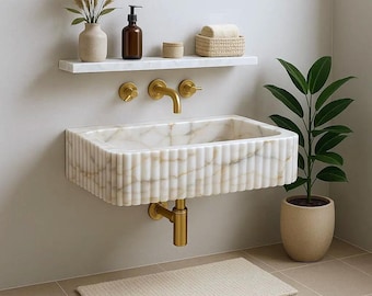 Puede incluir: Un lavabo de baño moderno de mármol blanco con detalles acanalados verticales. El lavabo tiene grifos dorados y está montado en la pared. Una estantería blanca encima contiene una botella marrón, un jarrón y una cesta tejida.
