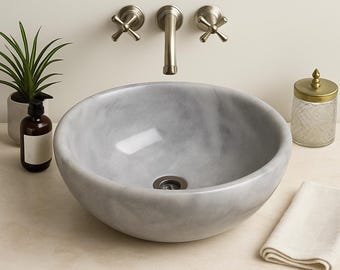 May include: A gray marble vessel sink with a brushed nickel faucet and cross handles. A small potted plant, a soap dispenser, a glass jar with a gold lid, and a folded white towel are on the countertop.