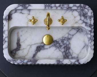 Può includere: Un lavandino rettangolare in marmo con bordo smerlato. Il lavandino presenta un motivo venato bianco e grigio. È dotato di rubinetteria color oro, tra cui un rubinetto, due maniglie a forma di croce e un copri-scarico. Il lavandino è posto su uno sfondo scuro.