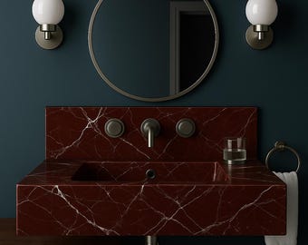 Puede incluir: Un lavabo de baño moderno de mármol rojo con vetas blancas. El lavabo tiene un grifo y un desagüe integrados, un vaso pequeño y un toallero. Un espejo redondo y dos apliques de pared con globos de luz están encima.