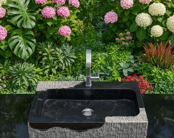 May include: A black rectangular sink with a silver faucet. The sink is made of dark stone and has a rough, textured edge. The background features lush greenery and pink and white flowers.