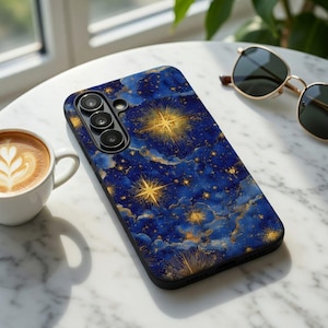Op de afbeelding: Een smartphonehoesje met een hemels ontwerp. De hoes heeft een donkerblauwe achtergrond met gouden sterren en wolkenpatronen. De telefoonhoes heeft een zwarte rand en een cameramodule met drie lenzen. Een kopje koffie en een zonnebril zijn ook zichtbaar.