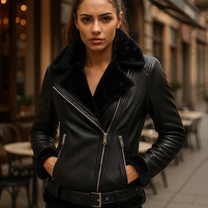 May include: A black leather jacket with a shearling collar and lining. The jacket features a belted waist, zippered pockets, and a front zipper closure. The model is wearing black jeans and has her hands in her pockets.