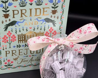 May include: A decorative Christmas ornament filled with rolled-up paper and a clear glass ball, tied with a pink floral ribbon. Beside it is a book titled "Pride and Prejudice" by Jane Austen, with a floral design on the cover.