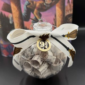 Peut inclure: Une boule de Noël en verre transparent remplie de papier roulé. L'ornement est décoré d'un ruban blanc avec des motifs de balais et d'un charm doré avec le texte "9 3/4 Kings Cross London".
