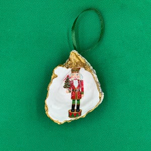 May include: A Christmas ornament featuring a painted nutcracker soldier inside a white oyster shell with gold trim. The nutcracker is red, green, and brown, holding a small green Christmas tree. A green ribbon is attached for hanging.