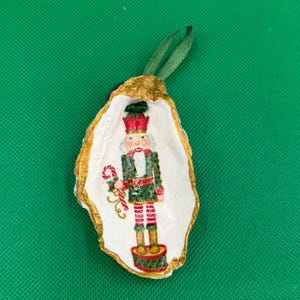 Puede incluir: Un adorno navideño con un soldado cascanueces pintado dentro de una concha de ostra blanca con un borde dorado. El cascanueces lleva un uniforme rojo y verde y sostiene un bastón de caramelo. Se adjunta una cinta verde para colgar.