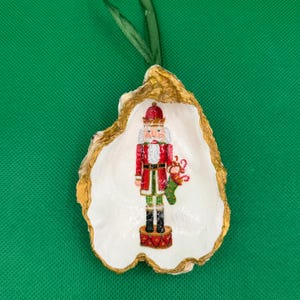 Puede incluir: Un adorno navideño con un soldado cascanueces pintado dentro de una concha de ostra blanca con un borde dorado. El cascanueces lleva un abrigo rojo y pantalones verdes, sosteniendo un calcetín. Se adjunta una cinta verde para colgar.