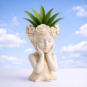 Peut inclure: Un pot de fleurs décoratif crème en forme de tête et d'épaules de femme, avec les yeux fermés et les mains touchant doucement son visage. La tête est ornée d'une couronne florale, et des feuilles vertes jaillissent du sommet. Des accents dorés rehaussent le design.