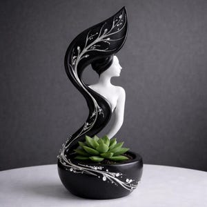 Op de afbeelding: Een zwart-witte decoratieve plantenbak met een silhouet van een vrouw en een vloeiend bladerontwerp. De plantenbak bevat een groene vetplant. Zilveren accenten en kristallen sieren het blad en de basis.