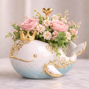 Peut inclure: Un vase en céramique en forme de baleine, peint en blanc avec une base bleu clair, orné d'accents dorés et de strass. Le vase est surmonté d'une couronne dorée et d'un fleuron en forme de cœur, et rempli de roses roses et de verdure.