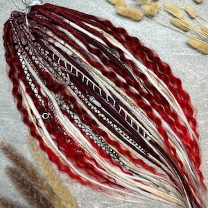 May include: A bundle of red, white, and burgundy dreadlocks, tied together with a silver ribbon. The dreadlocks are decorated with silver and black accents. The hair extensions are wavy and have a gradient of colors.