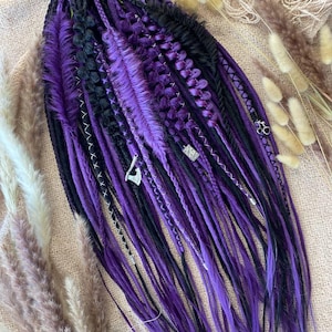 Puede incluir: Un conjunto de rastas moradas y negras con abalorios plateados decorativos y costuras blancas. Las rastas están dispuestas sobre una superficie de tono neutro, con hierbas ornamentales secas en el fondo. Las rastas tienen un efecto degradado, pasando del negro al morado.