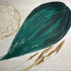 May include: A bundle of dark green and black synthetic hair braids, transitioning to a vibrant green at the ends. The braids are arranged in a teardrop shape, with a decorative doily and dried wheat stalks in the background.