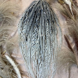 Può includere: Un set di dreadlock grigi e bianchi con varie trame, tra cui trecce e torsioni. I dreadlock sono disposti in un fascio e sono circondati da erbe ornamentali essiccate. La sfumatura di colore va dal grigio al bianco.