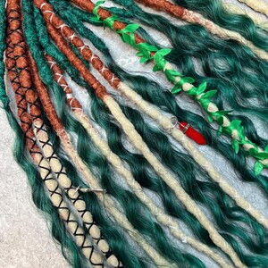 May include: A set of synthetic hair extensions featuring a mix of wavy and dreadlock textures in shades of forest green, copper brown, and cream. The strands are accented with black and white thread wraps, a green leaf vine, and a red crystal charm.