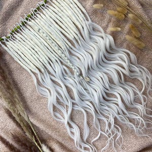 May include: White and silver dreadlocks with wavy ends, arranged on a textured, light brown surface. The dreadlocks are attached to a green base. Dried ornamental grasses are in the background.