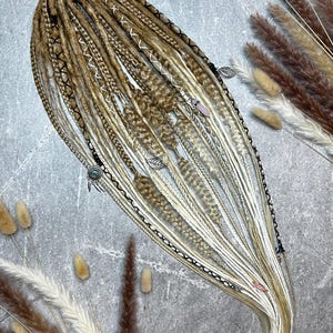 Könnte beinhalten: Ein Set aus blonden und braunen Kunst-Dreadlocks, verziert mit dekorativen Perlen und Charms. Die Dreads sind mit einem rosa Band zusammengebunden. Die Haarverlängerungen haben weiße, braune und schwarze Akzente.