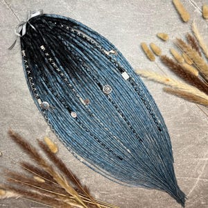 Op de afbeelding: Een set blauwe en zwarte dreadlocks met zilveren accenten en decoratieve bedels. De dreadlocks zijn vastgebonden met een zilveren lint. De haarverlengingen zijn gerangschikt op een grijze ondergrond met gedroogde planten.