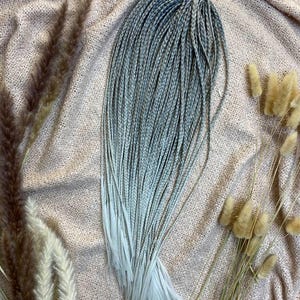 May include: A bundle of gray and white braided synthetic hair extensions. The braids are tightly woven and transition from a darker gray at the roots to a lighter, almost white, color at the ends. The hair is tied together with a purple band.