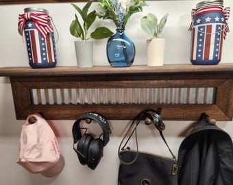 May include: A wooden shelf with a decorative metal insert, holding two mason jars painted with a patriotic design. The shelf also displays small potted plants and a blue glass vase. Below, a hat, headphones, a purse, and a jacket hang from hooks.
