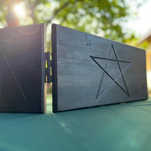 May include: Two-piece, dark grey wooden box with a pentagram carved into one side. The box is connected by a hinge, and the other side has a geometric design. The box is sitting on a green surface.