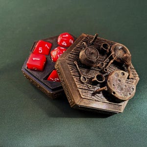 Op de afbeelding: Een gedetailleerde, zeshoekige dobbeldoos met een rustieke, steampunk esthetiek. De doos is open en onthult rode dobbelstenen. Het deksel heeft miniatuur keukengerei en een klok, allemaal in een bronzen afwerking.