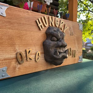Puede incluir: Diana de dardos de madera con las palabras "HUNGRY INN" y "OKC" en relieve. Una cara de orco tallada, de color oscuro, está en el centro. Las esquinas metálicas decorativas y los dardos completan el diseño.