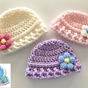 May include: Three handmade crochet baby hats in cream, pink, and lavender. Each hat features a decorative flower in contrasting colors. The hats are arranged on a white surface, showcasing the delicate craftsmanship.