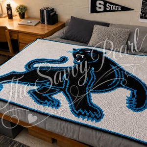 May include: A rectangular crochet blanket featuring a large black panther graphic with blue outlines on a white background. The panther is depicted in a pouncing position with its mouth open, showcasing white teeth. The blanket has a blue border.