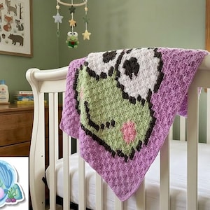Può includere: Una coperta per neonati lavorata all'uncinetto a mano con un personaggio rana verde pixelato, grandi occhi bianchi, pupille nere e guance rosa, su uno sfondo viola lavanda testurizzato, drappeggiata sul lato di una culla in legno bianco.