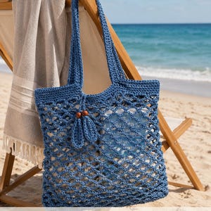 May include: A blue crochet tote bag featuring an open diamond mesh pattern, two long shoulder straps, and a decorative accent with two small blue crochet leaves attached to two round wooden beads. The text The Savvy Pearl is visible.