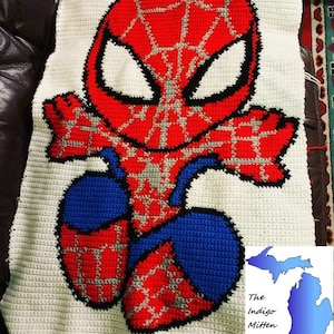 May include: A crocheted blanket featuring a cartoon depiction of Spider-Man. The superhero is rendered in red, blue, and gray, with a white background. The blanket is handmade and has a textured appearance.
