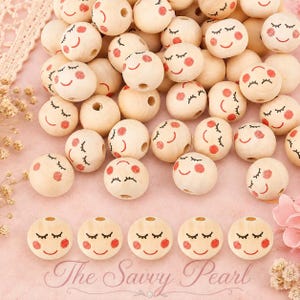 May include: A collection of round, light-colored wooden beads, each featuring a printed face with closed eyes, a simple curved smile, and pink circular cheeks. The beads are arranged in a pile with five additional beads lined up below.