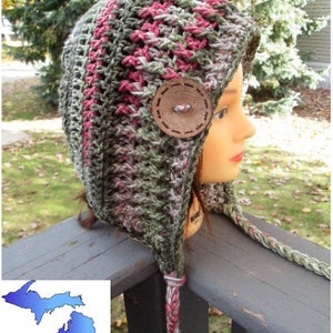 Puede incluir: Un gorro de ganchillo con orejeras que presenta un patrón de hilo jaspeado en tonos verde oliva, gris y magenta. El gorro está adornado con un botón de madera marrón grande y redondo en el lateral e incluye largas tiras de hilo trenzado.