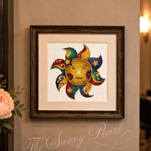 May include: A framed cross-stitch artwork featuring a central golden sun face surrounded by stylized rays in shades of red, blue, and yellow. The design includes small star motifs and is set against a white fabric background within a dark frame.