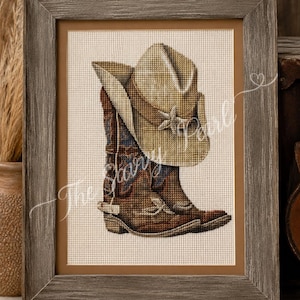 May include: A framed cross-stitch embroidery featuring a tan cowboy hat resting on a brown leather cowboy boot with blue accents. The artwork is displayed in a weathered gray wooden frame with a tan mat, showcasing detailed needlework stitches.