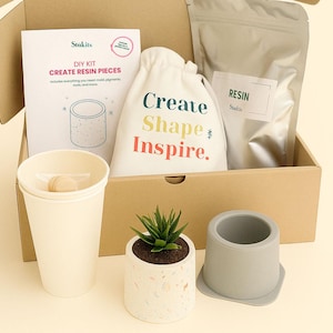 Może przedstawiać: Zestaw do samodzielnego wykonania elementów z żywicy w kartonowym pudełku. Zestaw zawiera broszurę, torbę z napisem "Create Shape Inspire", żywicę, kubek i małą roślinę sukulent w ozdobnej doniczce. W zestawie znajduje się również szara forma do żywicy.