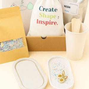 Puede incluir: Un kit de resina DIY con varios componentes. Incluye una bolsa con el texto "Create Shape Inspire", una bolsa de resina, una taza, palitos para remover y dos bandejas ovaladas. El kit también contiene una bolsa de confeti colorido.