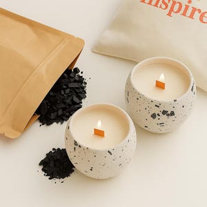 May include: Two lit, round candles in speckled white ceramic containers. A brown paper bag spills black charcoal pieces. A white bag with the word "inspire" in orange is in the background. The candles have wooden wicks.