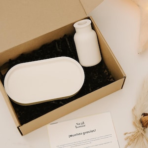 Puede incluir: Un jarrón de cerámica blanco y una bandeja ovalada blanca con borde dorado están dentro de una caja de cartón con papel negro. Una tarjeta con el texto "Still home ¡Muchas gracias!" también está en la caja.
